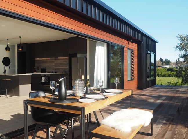 An outdoor wooden dining table set with plates and glasses on a patio next to a modern kitchen with black cabinets and a refrigerator at Pinot Longbox - Martinborough Holiday Home in Martinborough