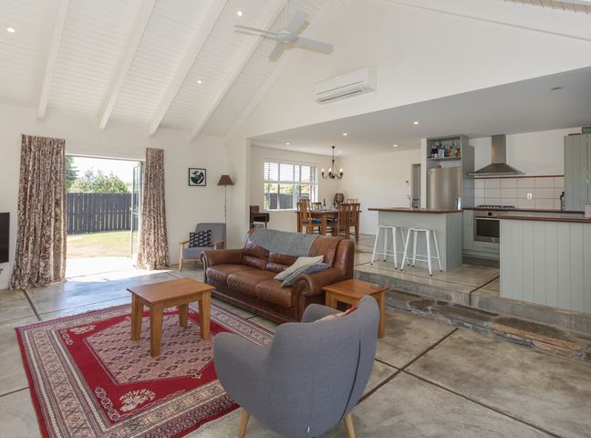 A living room with leather sofa gray armchairs wooden tables and red rug adjacent to an open kitchen and dining area at Modern Cottage Charm - Albert Town near Wanaka in Wanaka