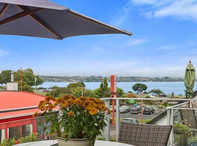 A balcony with a table and flowers overlooking a parking lot and a lake with a blue sky at Relax on Roberts Taupo Holiday Home Taupo Central