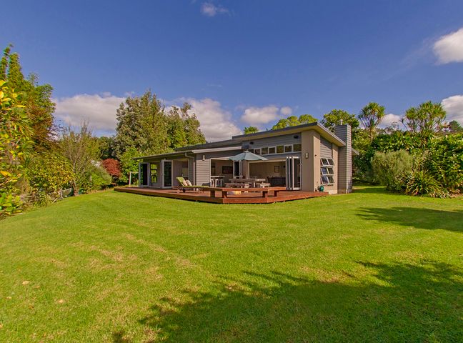 A house with a wooden deck, outdoor furniture, and green lawn surrounded by trees at Cooks Getaway - Cooks Beach Holiday Home in Cooks Beach