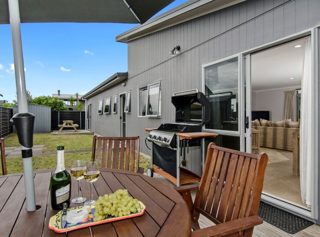 A patio with a wooden table and chairs a bottle of wine and grapes on the table a barbecue grill and an open sliding door showing a living room at Beauty on Bream Bay - Ruakaka Holiday Home in Ruakaka