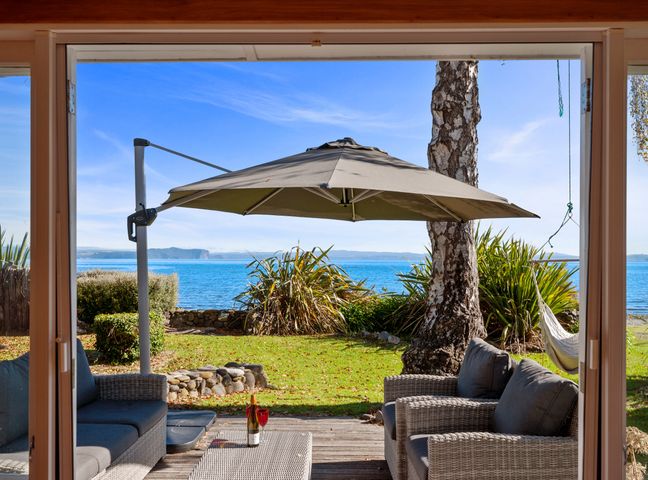 An outdoor patio with wicker sofas and a table under an umbrella overlooking a lake with plants and a hammock at North Cole Cottage - Hatepe Lake Front Home in Hatepe