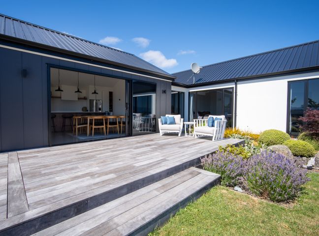 A wooden deck with two white armchairs and a small table outside a modern house with large sliding glass doors and garden bushes at Lake House - Te Anau Holiday Home in Te Anau