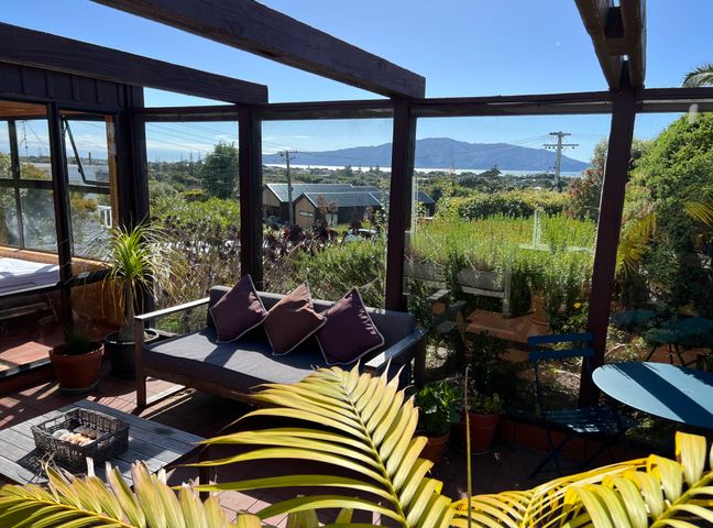 A sunroom with a sofa and cushions a small table and chair and plants with a view of a garden and mountains at Tranquility Base - Waikanae Beach Holiday Home in Waikanae Beach