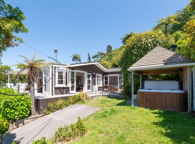 A backyard with a hot tub under a wooden pavilion and a house with open windows and doors at Kiwiana Bach - Lake Tarawera Holiday Home in Lake Tarawera