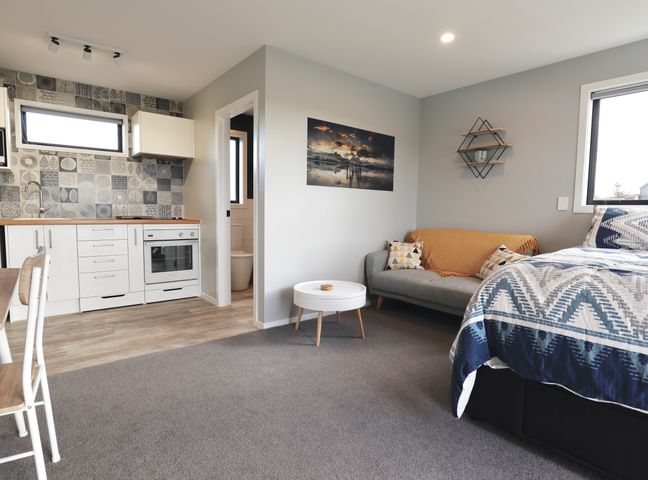 A studio room with a kitchenette, a gray sofa with pillows and a blanket, a white round table, a bed with patterned bedding and a wall photo at The Tui Studio - Te Anau Holiday Studio in Te Anau