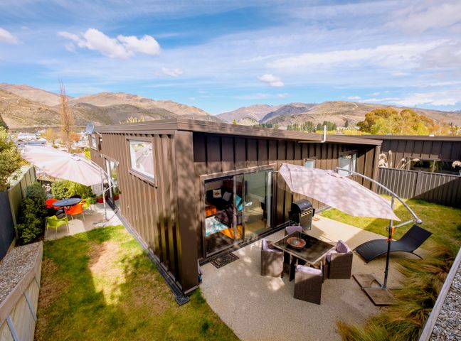 An outdoor patio area with chairs a table umbrellas and a barbecue next to a brown building with mountains in the background at Kawerau River Retreat - Cromwell Holiday Home in Cromwell