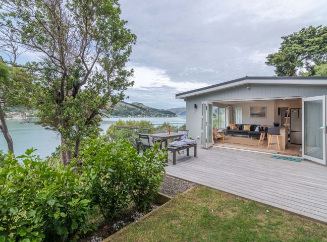 An outdoor deck with table and chairs next to a modern living room overlooking a body of water at Otahuahua - Akaroa Holiday Home in Akaroa