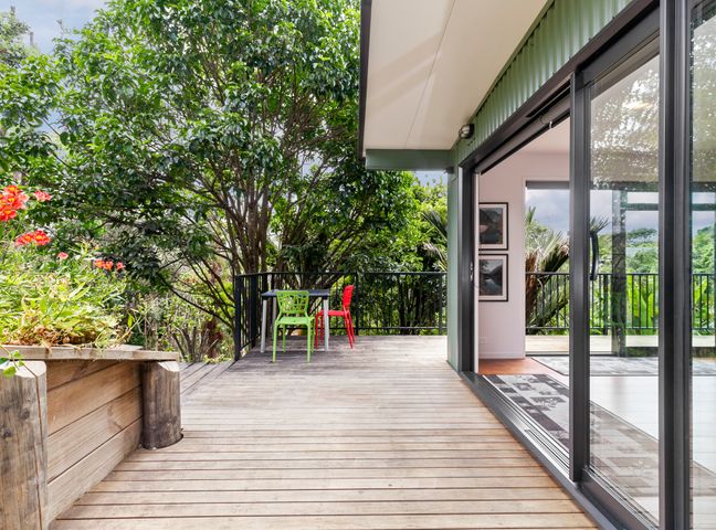 An outdoor wooden deck with two chairs and a table surrounded by trees and plants at Scotts Landing Bach Retreat Mahurangi East Algies Bay