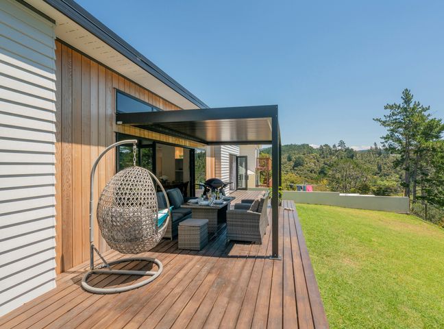 An outdoor wooden deck with a hanging chair seating area dining table and barbecue grill overlooking grass and trees at Treetop Bach - Whangamata Holiday Home Whangamata