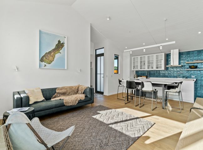 A living room with a dark sofa, white chair, rug, and a kitchen with bar stools and blue tiled backsplash at Bull Ridge Escape Wanaka