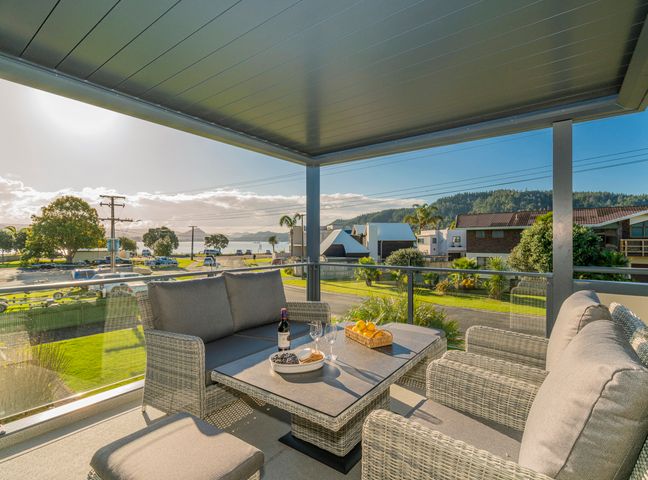 An outdoor covered seating area with wicker furniture and a table with wine and food overlooking a street and water at Harbour View - Whangamata Holiday Home in Whangamata