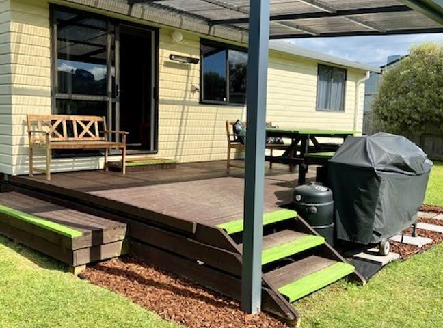 A wooden patio with a bench table grill and barbecue next to a light yellow house at Relax Cabin Waihi Beach