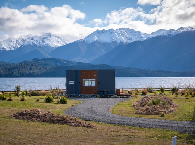 A small house with a gravel path and plants in front of a lake and mountains at Tiny Piece of Paradise - Te Anau Tiny House in Te Anau