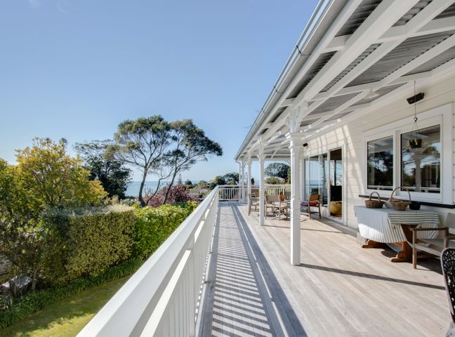 An outdoor balcony with wooden flooring white railings chairs and a covered roof overlooking trees and the sea at The Verandah - Napier Holiday Home in Bluff Hill