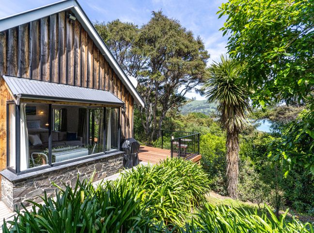 A house with wooden siding and a stone base next to a deck surrounded by trees and plants at Woodside - Akaroa Holiday Cottage in Akaroa