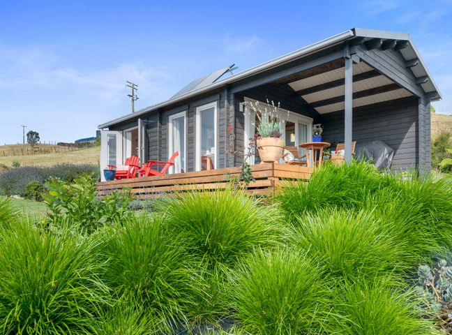 An outdoor view of a cabin with a wooden deck and red chairs surrounded by green plants at Kaitipu - Papamoa Hills Holiday Cabin in Papamoa