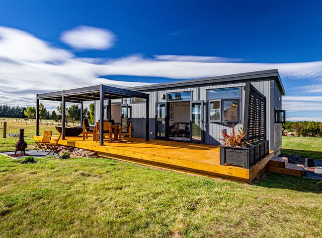 A small gray house with a wooden deck a pergola with chairs and table and a grassy yard at Mangateitei Views  Rangataua
