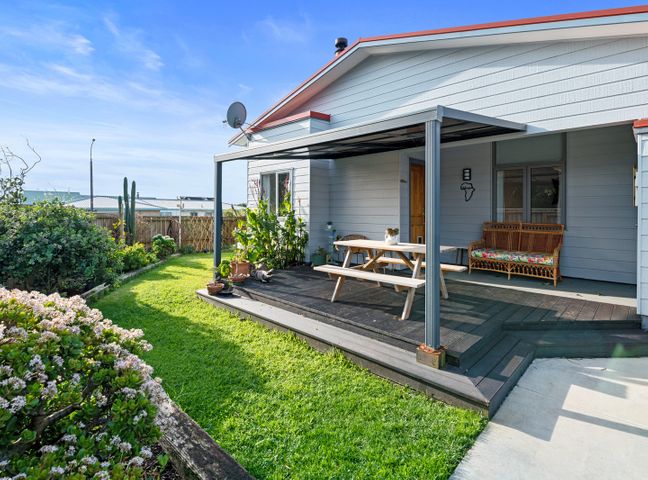 A backyard with a covered wooden deck table and a wicker bench surrounded by grass and plants at Ekhaya - New Plymouth Holiday Home in New Plymouth