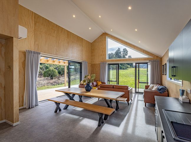 A living room with wooden dining table and benches brown sofas and large windows showing greenery at Manawa - Ohakune Holiday Home in Ohakune