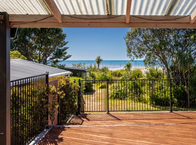 A wooden deck with metal railing overlooking a grassy garden with trees and a beach in the background at Godwit Cottage - Pakawau Beachfront Holiday Home Pakawau