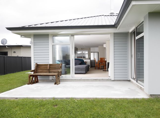 A patio area with a wooden bench and sliding glass doors leading to a living room with a gray sofa and wooden dining chairs at Fiordland Getaway - Te Anau Holiday Home in Fiordland