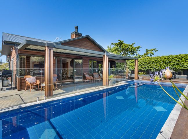 An outdoor swimming pool next to a wooden house with a glass fence and patio chairs at Hikanui Haven - Havelock North Holiday Home in Havelock North
