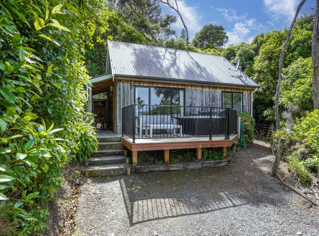 A small wooden cottage with a metal roof and a deck surrounded by greenery at Bushside Cottage - Akaroa Holiday Home in Akaroa