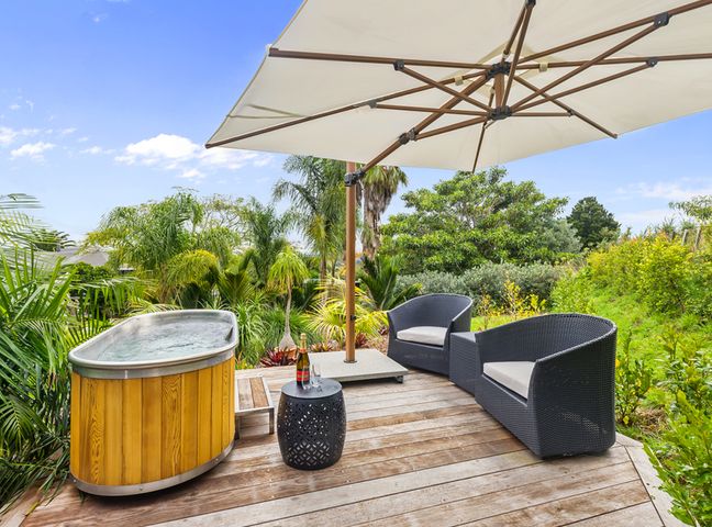An outdoor wooden deck area with a hot tub two black wicker chairs a black side table with a bottle and glasses and a large white umbrella surrounded by greenery at Country Idyll - Sandspit Holiday Cottage in Sandspit