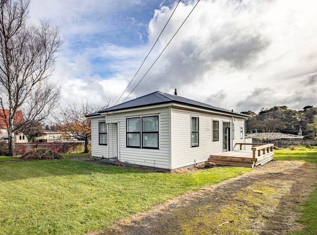 A small white house with multiple windows and a wooden deck surrounded by grass and trees at Rata Retreat - Ohakune Holiday Home in Ohakune