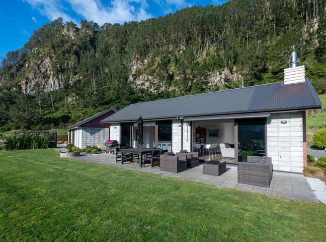 A single-story house with outdoor seating and dining area on a patio with a forested cliff in the background at Lakehouse Retreat - Whakamaru Holiday Home Waipamu Station 45km from Taupo
