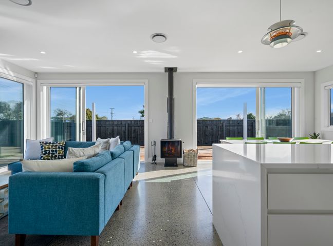 A living area with a blue sofa, wood stove, large windows, and a white kitchen island at Stylish Serenity - Martinborough Holiday Home in Martinborough