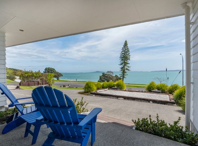 A covered patio with blue chairs overlooking the ocean and a road with trees and shrubs at Maraetai Couples Retreat - Omana Beach Apartment in Maraetai