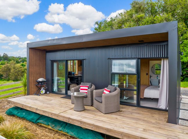 A wooden deck with two wicker chairs and a small table outside a modern cabin with sliding glass doors and a grill nearby at Kuku Kabin - Waimate North Eco Holiday Cabin in Waimate North 16km from Kerikeri 24km to Paihia