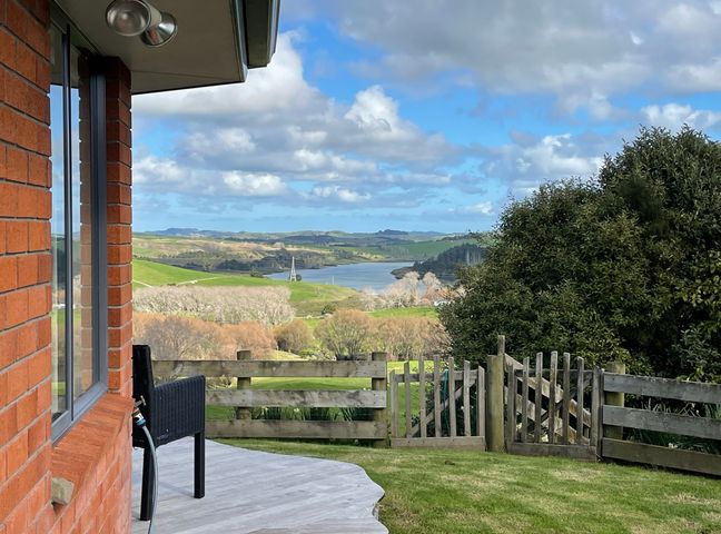 A view of a river and hills from a wooden deck outside a red brick house with a black chair and a wooden fence with a gate at Kaiwaka Paradise Kaiwaka