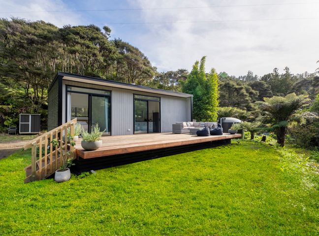 An outdoor wooden deck with seating and bean bags attached to a small modern cabin surrounded by grass and trees at Valley Hideaway - Kuaotunu Holiday Cabin in Kuaotunu