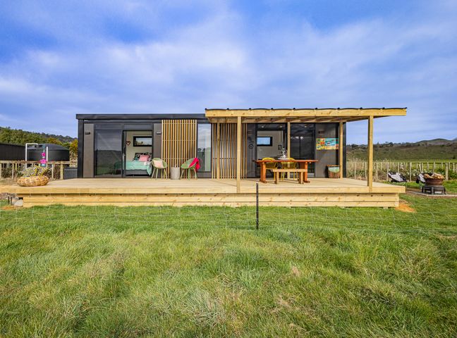 A small modern house with a wooden deck and outdoor dining furniture in a grassy field at Whare Awa - River House in the Rangitikei