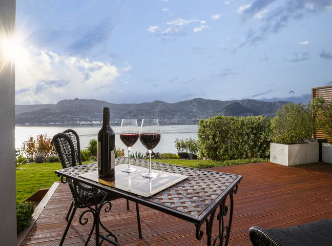 An outdoor patio with a metal table holding two glasses of red wine and a bottle overlooking water and mountains at Sanctuary At The Bay - Diamond Harbour Retreat Diamond Harbour