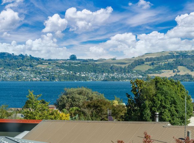 A view of a lake with houses on hills in the background and trees and rooftops in the foreground at Poppy's Bach - Lake Taupo Holiday Home in Waipahihi