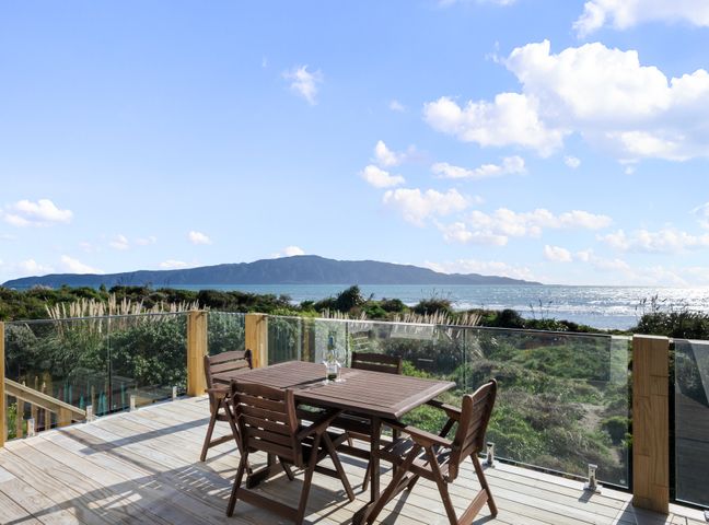 A wooden deck with a table and four chairs overlooking greenery and the ocean at Sunset Shores Paraparaumu Beach