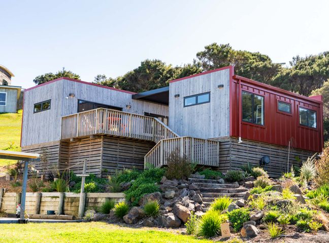 A modern raised house with wooden and red siding on a hillside with stone steps and greenery at Whero Pods - Mangawhai Heads Holiday Home in Mangawhai Heads