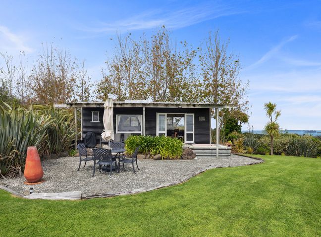 A small black cabin with a white roof surrounded by trees and bushes with a gravel patio and outdoor table with chairs at Tawharanui Cabin - Rural Cabin Studio in Tawharanui