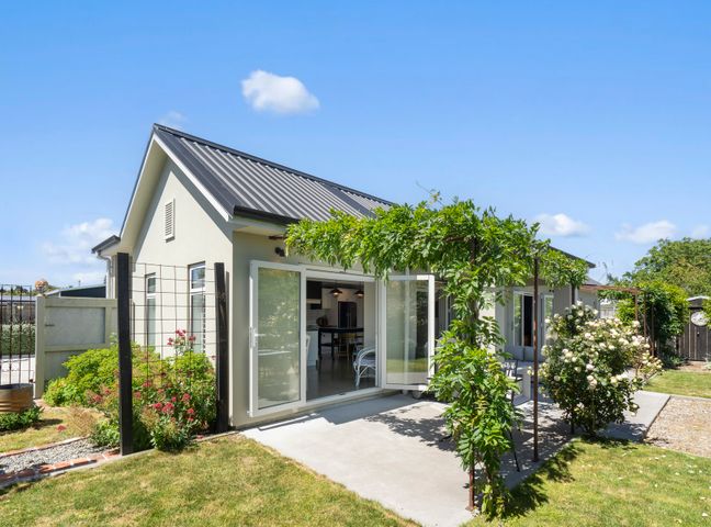 An exterior view of a house with open patio doors and a garden with green plants and flowers at Provence on Cologne - Martinborough Holiday Home in Martinborough