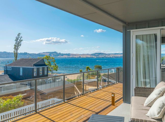 A balcony with a glass railing and outdoor furniture overlooking a lake and houses at Willow Lake House - Lake Taupo Holiday Home in Rainbow Point