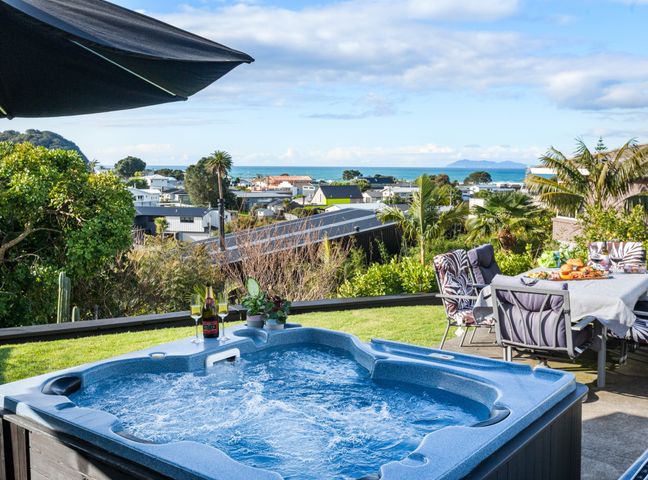 An outdoor area with a hot tub pool champagne bottle and glasses a dining table with chairs and a view of houses and the sea at Two Palms - Waihi Beach Holiday Apartment in Waihi Beach