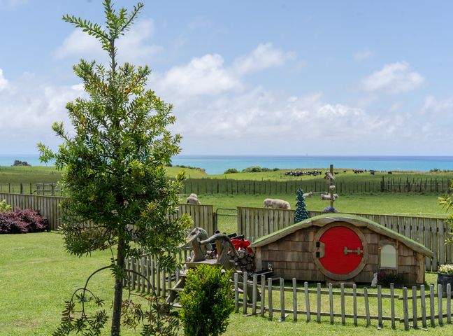 A garden with a small wooden shed with a red circular door and fenced area with animals grazing near the coast at Onaeros Cute Little Cottages - Taranaki Retreat in Waitara