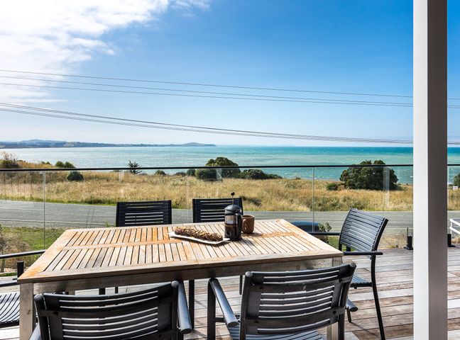 An outdoor wooden table with black chairs on a deck overlooking a grassy area and a body of water at Moeraki Beach Haven - Otago Beachfront Home in Otago
