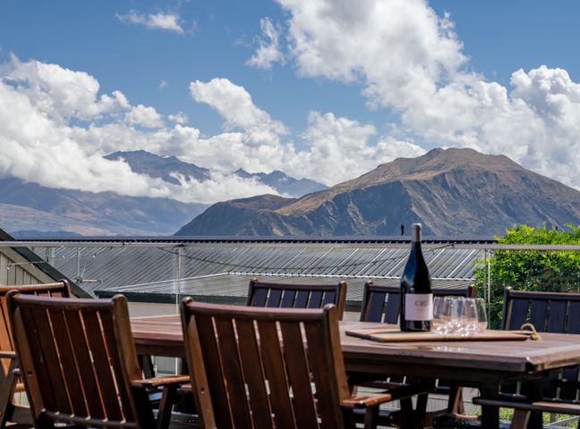 An outdoor dining area with wooden chairs and table with a wine bottle and glasses with mountains and clouds in the background at Peak Retreat Wanaka Holiday Home in Wanaka