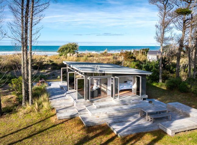 A small house with large glass doors opening to a wooden deck with picnic table near the beach at Mahanga Dunes Retreat - Mahia Holiday Home in Mahia Peninsula