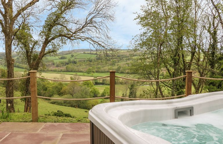 An outdoor hot tub on a stone patio overlooking a fenced green countryside at Little Silver Leaf in High Bickington